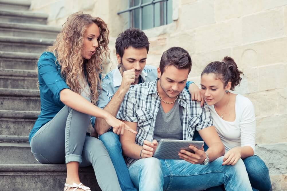 Vier Freunde sitzen auf einer Treppe und schauen gemeinsam auf einem Tablet, wie man Lotto mit System spielt