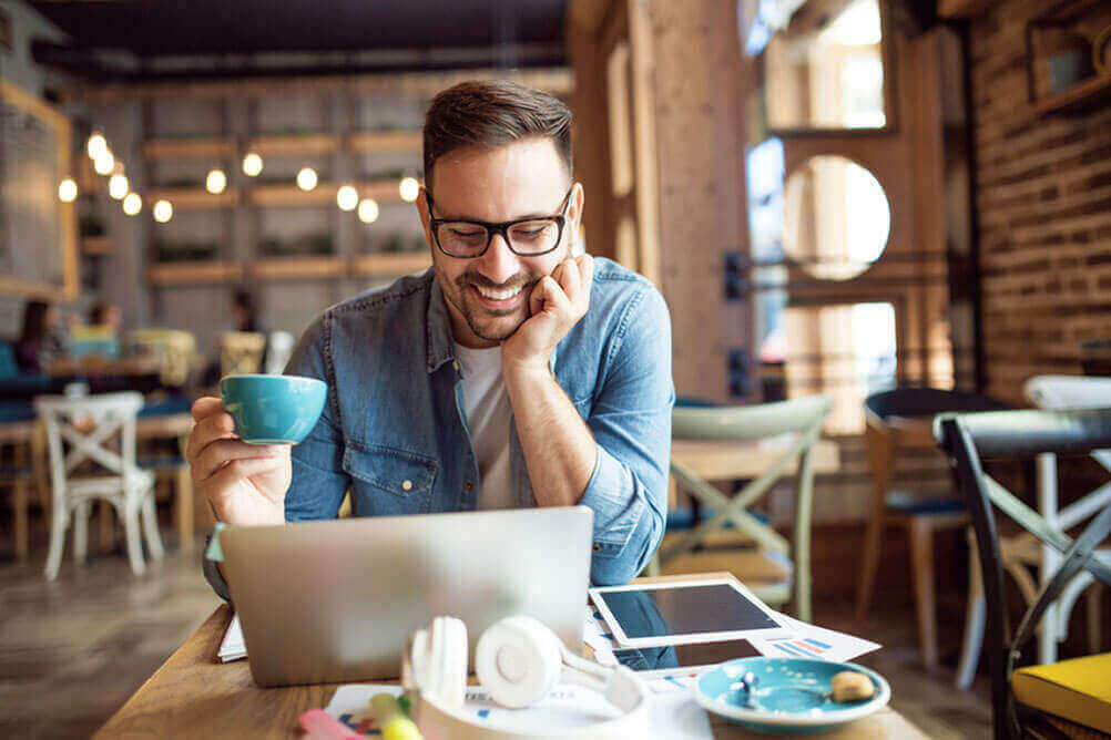Mann sitzt lächelnd an seinem Laptop und trinkt Kaffee, während er Guthaben auf sein Spielerkonto im Lottoland einzahlt
