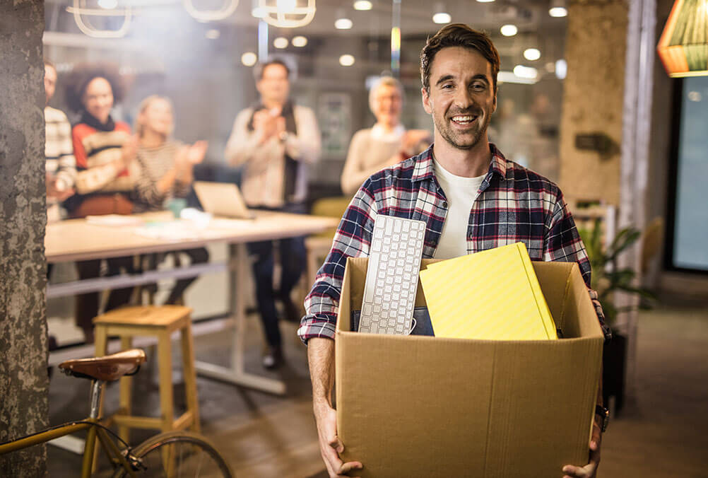 Ein lächelnder Mann trägt einen Karton mit seinem Besitz und verlässt seinen Arbeitsplatz